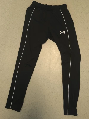 Svarta träningsbyxor från Under Armour - Säljer ett par svarta träningsbyxor från Under Armour med vita detaljer längs benen och logga på låret. Byxorna har resår i midjan med dragsko och är tillverkade i ett lätt och stretchigt material som passar perfekt för träning.