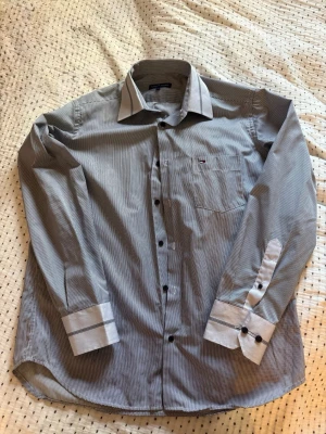Randig skjorta från Tommy Hilfiger - Snygg randig skjorta från Tommy Hilfiger i grå/silver. Klassisk krage, långa ärmar och knappar framtill samt på manschetterna. Bröstficka med liten logga. Perfekt till jeans eller chinos för en clean look.
