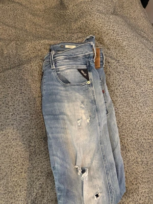 Blå slitna jeans från Replay - Säljer ett par ljusblå jeans från Replay med slitningar och hål på benen. Jeansen har klassisk femficksmodell och raka ben. Materialet är denim i bomull med en snygg tvättad look och detaljerade slitningar för en trendig stil.