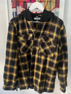 Gul och svart rutig overshirt Bershka - Overshirt från Bershka i svart och gul rutig design. Skjortjackan har tryckknappar, två bröstfickor med lock och en svart huva i sweatshirtmaterial. Perfekt lager på lager-plagg med avslappnad passform och trendig streetkänsla. Snöret i luvan saknas enligt bild men går enkelt att ersätta med eget snöre.