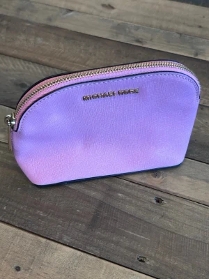 Rosa necessär från Michael Kors - Snygg necessär i rosa från Michael Kors med guldfärgad dragkedja och logga framtill. Tillverkad i ett tåligt, strukturerat material med rundad form och rymligt fack. Perfekt för smink eller småsaker i väskan.