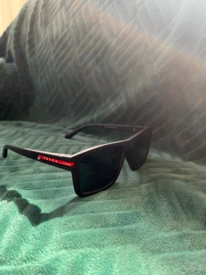 Svarta solglasögon från Prada - Snygga svarta solglasögon från Prada med breda bågar och rektangulär form. Diskret logga på sidan och en röd detalj längs skalmen ger en sportig touch. Perfekta för dig som vill ha en stilren och modern look. PRISET ÄR EJ HUGGET I STEN