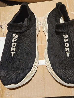 Svarta slip-on sneakers med text - Svarta slip-on sneakers med vit sula och texten 'SPORT' i vitt på ovansidan. Skorna har en stickad ovandel i textil som ger en strumpliknande passform och en chunky, räfflad sula i gummi. Perfekta för en sportig och avslappnad stil.