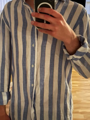 Randig linneskjorta från Massimo Dutti - Snygg skjorta i 100% linne från Massimo Dutti med breda blå och vita ränder. Skjortan har klassisk krage, knäppning framtill och långa ärmar. Perfekt för en avslappnad och stilren look.