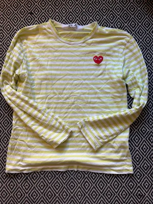 Randig långärmad t-shirt från Play - Gul och vit randig långärmad t-shirt från Play Comme des Garçons med det klassiska röda hjärtat broderat på bröstet. Skön passform och tillverkad i mjuk bomull. Perfekt för dig som gillar streetstyle och vill sticka ut lite ex