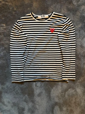 Comme des Garçons Tröja - Säljer en svartvit randig långärmad topp från Comme des Garçons Play med det ikoniska röda hjärtat broderat på bröstet. Klassisk rund halsringning och mjuk bomullskvalitet. Perfekt för dig som gillar streetstyle och vill ha något tidlöst men ändå lekfullt.