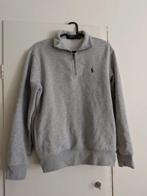 Grå halvzip sweatshirt Polo Ralph Lauren - Klassisk grå sweatshirt med halv dragkedja från Polo Ralph Lauren. Tröjan har hög krage, ribbade muddar och den ikoniska blå loggan broderad på bröstet. Perfekt för en avslappnad och stilren look.