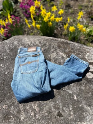 Nudie Jeans ”Lean Dean” - Säljer ett par klassiska ljusblå jeans från Nudie med fem fickor och snygga slitningar. Jeansen har raka ben och detaljerade sömmar, samt en brun läderpatch med logga bak i midjan. Perfekta för en avslappnad och trendig look.