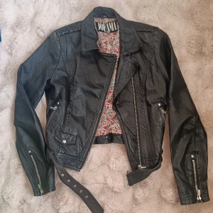 Svart skinnjacka Jofama by Kenza - Snygg svart skinnjacka från Jofama by Kenza med coola detaljer som dragkedjor vid ärmslut, bälte i midjan och blommigt innerfoder. Klassisk bikerstil med snedställd dragkedja och axeldekorationer. Perfekt för dig som vill ha en edgy look. Strl 34. Katt finns i hemmet. Rökfritt.