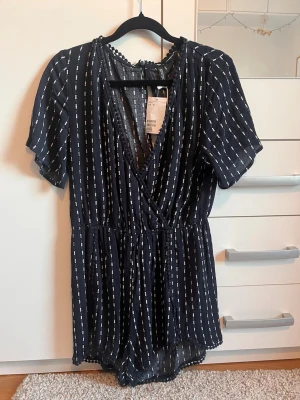 Mörkblå randig jumpsuit från Divided - Snygg mörkblå jumpsuit från Divided med vita vertikala ränder och korta ärmar. Djup v-ringning och resår i midjan för en skön passform. Tillverkad i ett lätt och luftigt material, perfekt för varma dagar.