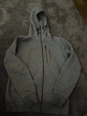 Grå zip-hoodie från Peak Performance - Snygg grå hoodie med dragkedja från Peak Performance. Har huva med dragsko och en bröstficka med dragkedja. Perfekt för chill dagar eller när du vill ha en enkel och stilren look. Har ett stygn vid sidan av fickan med det är inget som märks 💕storleken är i xs men känns som S 