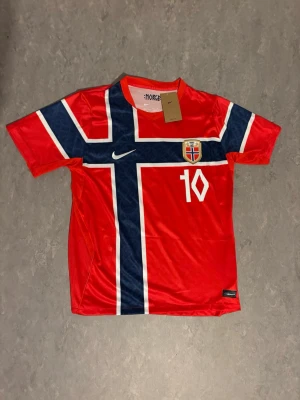 Norge fotbollströja - Röd Norge fotbollströja från Nike med stort norskt kors i blått och vitt, nummer 10 och Ødegaard på ryggen. Tröjan har korta ärmar, rund hals och klubbmärke på bröstet. 