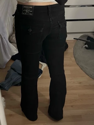 Svarta bootcut-jeans från Wrangler - Säljer ett par svarta bootcut-jeans från Wrangler med klassiska fickor och snygga detaljer på bakfickorna. Jeansen har hög midja och är tillverkade i ett stretchigt denimtyg som sitter bekvämt. Perfekta för en trendig och avslappnad look.