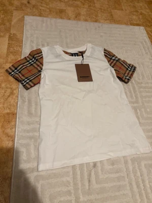 Vit Burberry t-shirt med rutiga ärmar - Snygg vit t-shirt från Burberry med klassiska rutiga korta ärmar i beige, svart och rött. T-shirten har rund halsringning och är tillverkad i mjuk bomull. Perfekt för en trendig och stilren look.