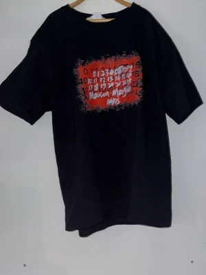 Maison Margiela Paris svart t-shirt - Svart t-shirt från Maison Margiela Paris med tryck i rött och vitt framtill. Passar en runt 165-170
