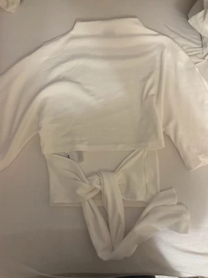 Tee back tie från Gina tricot  - Trendig vit croppad topp med knytband i midjan och trekvartsärm. Toppens material känns mjukt och stretchigt, perfekt för en stilren look. Enkel att matcha med både jeans och kjol för en fräsch outfit.