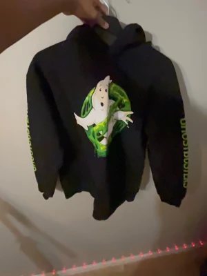 Svart Ghostbusters hoodie - Svart hoodie med stort Ghostbusters-tryck på bröstet och neongrön text 'GHOSTBUSTERS' längs båda ärmarna. Klassisk huva och ribbade muddar. Perfekt för dig som gillar retro och popkultur.