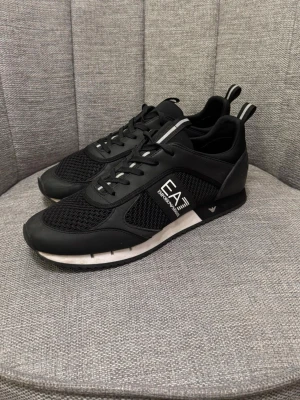Svarta EA7 Emporio Armani sneakers - Snygga svarta sneakers från EA7 Emporio Armani med vit logga på sidan. Skorna har meshpaneler för extra andningsförmåga, svarta snören och vita detaljer på sulan. Perfekta för en sportig och trendig look.