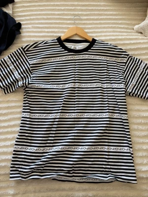 Randig svartvit t-shirt från Polar - Säljer en svartvit randig t-shirt från Polar med logotyptryck i rader över hela tröjan. T-shirten har svart krage och är tillverkad i mjuk bomull. 