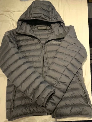 Grå dunjacka med huva Peak Performance - Snygg grå dunjacka från Peak Performance med huva och dragkedja framtill. Jackan har två sidofickor med dragkedja och quiltad design som håller värmen. Perfekt för kalla vinterdagar och enkel att matcha med olika outfits.
