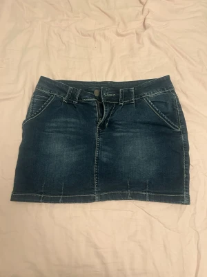 Mörkblå jeanskjol med fickor - Snygg mörkblå jeanskjol med klassiska fickor fram och bak, bälteshällor och knappstängning. Kjolen har en kort passform och tydliga sömmar som ger en cool denimlook. Perfekt att matcha med en t-shirt eller hoodie.