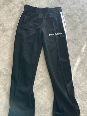 Palm Angels svarta mjukisbyxor - Svarta mjukisbyxor från Palm Angels med vita sidoränder och logga på vänster ben. Byxorna har resår i midjan och raka ben. Tillverkade i ett glansigt, sportigt material som känns riktigt skönt och avslappnat.  Lappen på bild 5 har lossnat på ena sidan men inget som märks av 