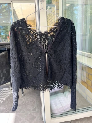 Svart spetsblus med v-ringning - Snygg svart blus i spets med v-ringning och långa ärmar. Blusen är croppad och har fina spetsdetaljer både fram och bak. Perfekt för dig som vill ha en trendig och elegant topp med unik känsla.