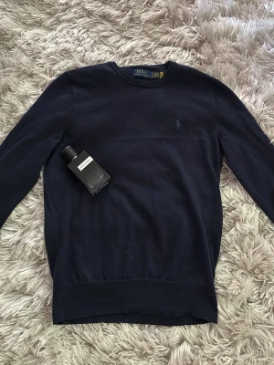 Mörkblå stickad tröja från Ralph Lauren - Mörkblå stickad tröja från Ralph Lauren med diskret broderad logga på bröstet. Tröjan har rund halsringning, långa ärmar och ribbade muddar. Perfekt för lager-på-lager och en klassisk stil.