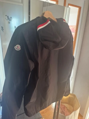 Svart Moncler vindjacka med huva - Svart vindjacka från Moncler med huva och rödvit randdetalj på huvan. Jackan har Moncler-logga på vänster ärm och är tillverkad i ett lätt, vindtätt material. Perfekt för blåsiga dagar och enkel att matcha med olika outfits.