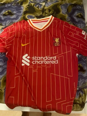 Liverpool FC röd matchtröja Nike - Säljer en röd Liverpool FC matchtröja från Nike med gula och vita detaljer. Tröjan har klubbmärke och sponsorlogga på bröstet samt ett unikt mönster i gult över hela plagget. Perfekt för fotbollsfans som vill visa sitt stöd på läktaren eller planen.
