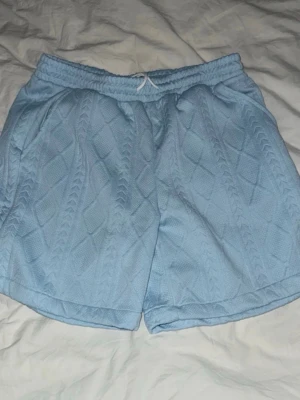 Ljusblå stickade shorts med snörning - Säljer ett par ljusblå stickade shorts med snyggt mönster och resår i midjan. Shortsen har snörning framtill och en avslappnad passform, perfekta för sommarens chill dagar.