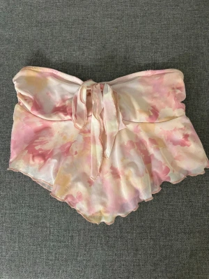 Pastellfärgad strapless topp med knyt - Supersöt topp i pastellrosa, gul och vit med knyt framtill. Toppen har ett flowigt och lätt tyg som ger en luftig känsla, perfekt för varmare dagar. Den har en mjuk och feminin look med oregelbunden fåll och romantiskt mönster. Inte säker på storleken men skulle vilja säga small