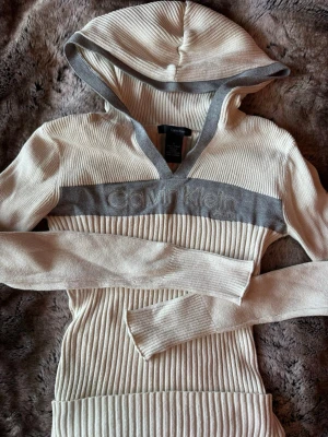 Beige ribbad hoodie från Calvin Klein - Snygg ribbad hoodie från Calvin Klein i beige med gråa detaljer och broderad logga över bröstet. Tröjan har långa ärmar, huva och V-ringning. Perfekt för en avslappnad och trendig look.