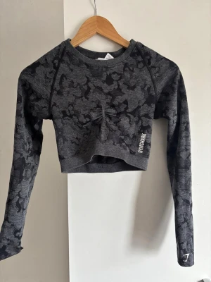 Grå cropped träningströja Gymshark - Cropped långärmad träningströja från Gymshark i mörkgrått camo-mönster. Tröjan har rynkdetalj framtill, tumhål i ärmsluten och en snygg cut-out i ryggen. Perfekt för gymmet eller annan träning.