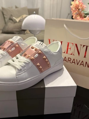 Valentino vita sneakers med rosa nitar - Snygga vita sneakers från Valentino med bred rosa rem över vristen dekorerad med ikoniska pyramidformade nitar i matchande rosa. Skorna har rund tå, vita skosnören och är tillverkade i slätt läder. Perfekta för dig som vill sticka ut med en lyxig touch.