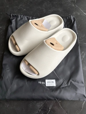 Beige Yeezy ”slides” YS-01  - Säljer ett par stilrena beigea slides från YS-01. Tofflorna har en chunky, räfflad sula och är tillverkade i ett mjukt, skummaterial som ger grym komfort. Perfekta att glida runt i hemma eller ute på stan. Enkel design med öppen tå och bred rem över foten. Kom med bud