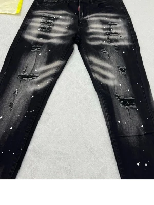 Svarta slitna jeans med färgstänk - Säljer ett par svarta jeans med coola slitningar och vita färgstänk. Jeansen har en smal passform och tydliga slitdetaljer på både fram- och baksida. Perfekta för dig som gillar en edgy och trendig stil. Materialet är klassisk denim.