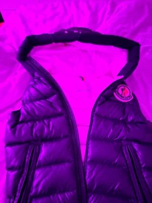 Mörkblå dunväst från Moncler - Snygg mörkblå dunväst från Moncler med klassisk logga på bröstet och två dragkedjefickor. Västen har en hög krage och quiltad design som ger en sportig och trendig look. Perfekt att slänga över en hoodie eller tröja för extra värme.