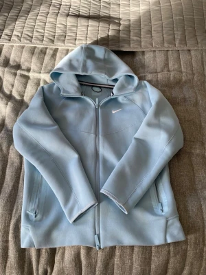 Nocta Nike hoodie - Snygg ljusblå hoodie från Nike med dragkedja framtill och två sidofickor med dragkedja. Klassisk huva och diskret Nike-logga på bröstet. Perfekt för en avslappnad och sportig stil. Tillverkad i mjukt och bekvämt material.