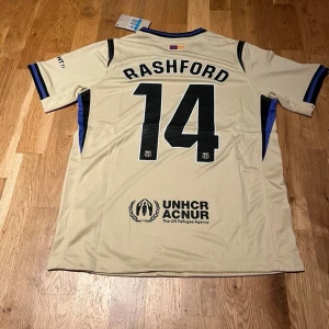 Barcelona away kit Rashford #14 - Snygg beige fotbollströja från FC Barcelona med Rashford och nummer 14 på ryggen. Tröjan har svarta och blå detaljer, UNHCR-logga, Spotify-logga på bröstet och klubbmärke. Tillverkad i lätt och ventilerande material, perfekt för träning eller match.