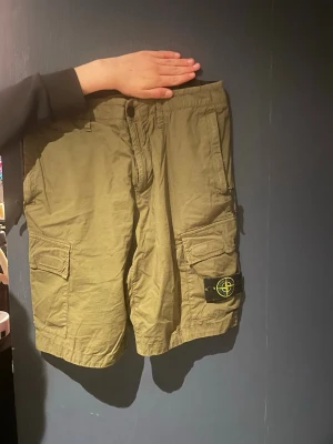 Beiga cargoshorts från Stone Island - Säljer ett par beiga cargoshorts från Stone Island med klassisk logotyp-patch på benet. Shortsen har flera fickor med lock och knappstängning samt en avslappnad passform. Perfekta för sommaren och streetwear-stilen.