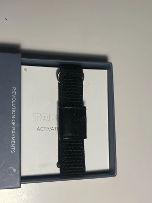Tapster svart armband för smartklocka - Snyggt svart armband från Tapster för smartklocka. Tillverkat i slitstarkt tygmaterial med metallspänne och diskret logga på lädret. Perfekt för dig som vill ha en stilren och funktionell wearable-accessoar.