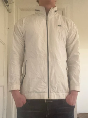 Vit vindjacka från Lacoste - Unik vit vindjacka från Lacoste med hög krage och dragkedja framtill. Jackan har diskret Lacoste-logga på bröstet. Tillverkad i lätt och vindtätt material, perfekt för blåsiga dagar.