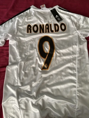 Real Madrid Ronaldo 9 matchtröja - Vit Real Madrid matchtröja från Adidas med Ronaldo och nummer 9 på ryggen. Tröjan har svarta detaljer, Siemens Mobile-tryck på framsidan och klubbmärke på bröstet. Tillverkad i glansigt funktionsmaterial, perfekt för fotbollsträning eller samling.