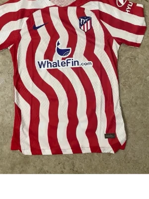 Atletico Madrid matchtröja Nike röd/vit - Säljer en Atletico Madrid matchtröja från Nike med röda och vita vågiga ränder, klubbmärke på bröstet och sponsorloggor. Tröjan har korta ärmar och är tillverkad i lätt och ventilerande polyester, perfekt för fotbollsträning eller match.