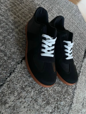 Svarta sneakers med brun sula - Stilrena svarta sneakers med vit snörning och brun gummisula. Skorna har en klassisk låg profil och är tillverkade i en mix av läder och textil. Perfekta för dig som gillar en clean och tidlös look. Ett par offbrand sneakers som ser superbra ut. Aldrig använda