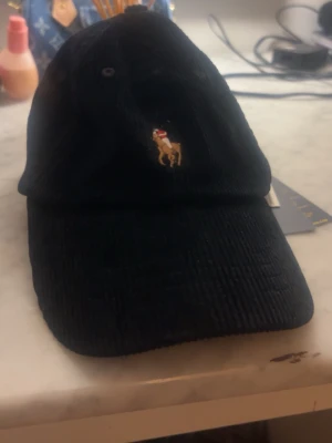 Svart keps från Polo Ralph Lauren - Snygg svart keps i manchestertyg från Polo Ralph Lauren med broderad logga framtill och justerbart spänne bak. Klassisk böjd skärm och broderad text baktill. Perfekt accessoar för en cool och avslappnad stil.