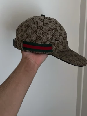 Gucci keps med GG-mönster - Snygg keps från Gucci i beige med klassiskt GG-monogram över hela kepsen. Har en grön och röd rand på sidan som ger den en ikonisk look. Justerbar rem baktill för perfekt passform. Materialet känns lyxigt och är typiskt för Gucci.