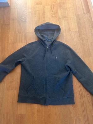 Mörkgrå hoodie från Polo Ralph Lauren - Mörkgrå hoodie med dragkedja från Polo Ralph Lauren. Klassisk design med huva och dragsko, två fickor framtill och den ikoniska lilla broderade loggan på bröstet. Perfekt för en chill och stilren look.