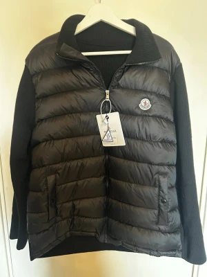 Svart Moncler cardigan - Snygg svart cardigan från Moncler med stickade ärmar och krage. Västen har två fickor framtill med knappstängning, dragkedja och Moncler-logga på bröstet. Passar även som L. Priset är inte hugget i sten🤩🙌